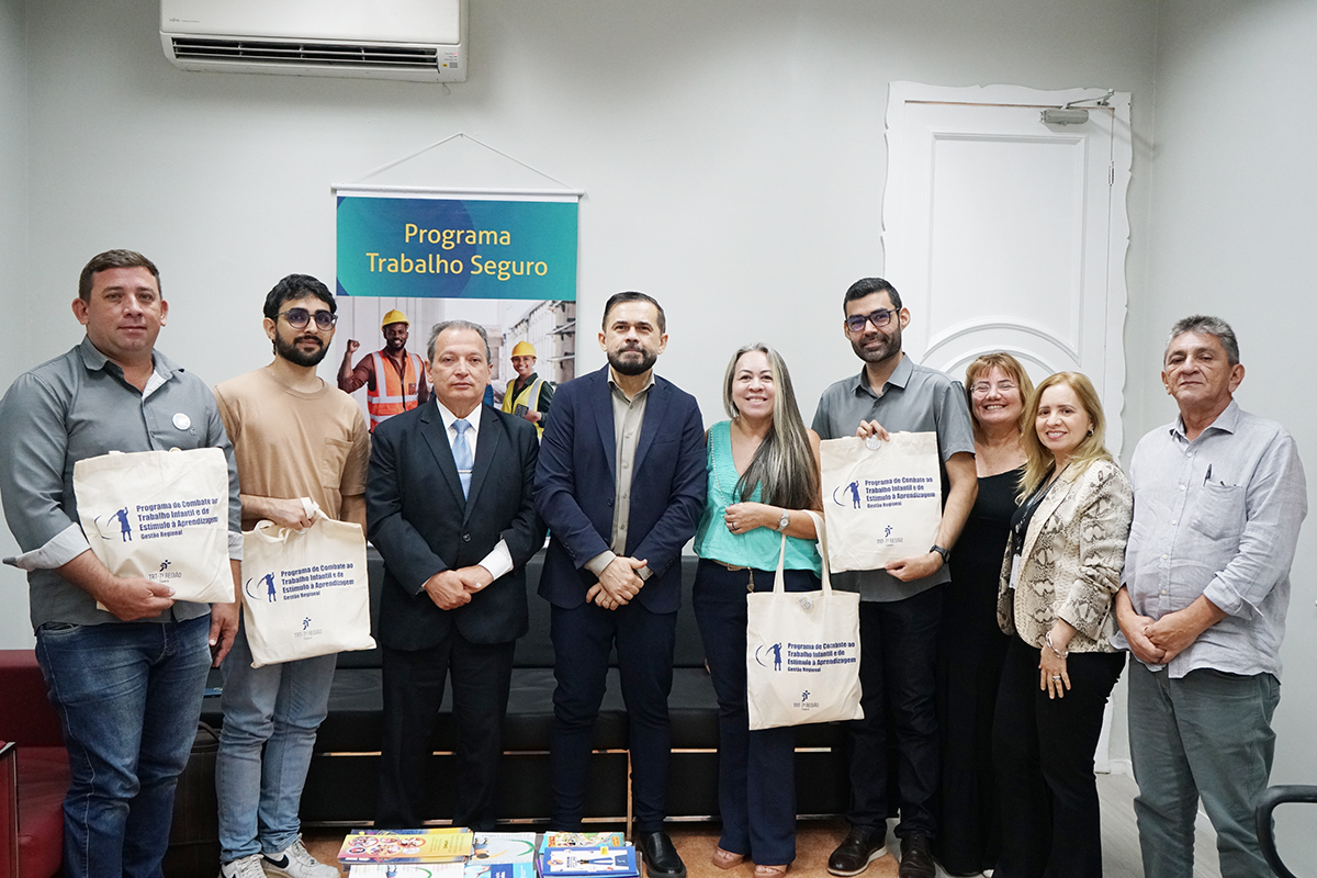 Grupo de nove pessoas posa alinhado em uma sala de escritório. Quatro delas seguram sacolas ecológicas brancas com a inscrição "Programa de Combate ao Trabalho Infantil e de Estímulo à Aprendizagem". Ao fundo, há uma porta branca ornamentada e um banner institucional com o título "Programa Trabalho Seguro".