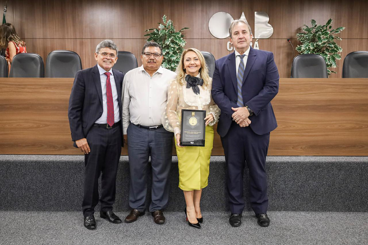 Quatro pessoas posam sorridentes à frente de uma bancada de madeira com o logotipo "OAB CEARÁ" ao fundo. No centro, uma mulher loira em blusa de tule branca e saia amarela segura uma placa de homenagem escura. Ela é ladeada por três homens em trajes formais (ternos e camisa social), em um auditório com poltronas pretas.