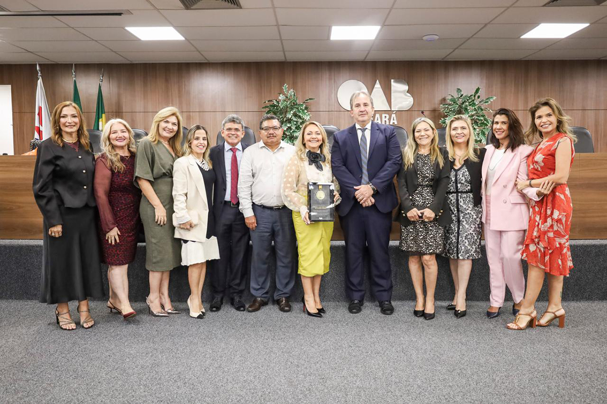 Um grupo de doze pessoas posa para uma foto oficial em um auditório da OAB Ceará. No centro, a mesma mulher da primeira foto segura sua placa, cercada por colegas homens e mulheres elegantemente vestidos em tons de preto, rosa, verde e vermelho. O grupo está posicionado à frente da bancada de madeira e de uma parede com plantas ornamentais.