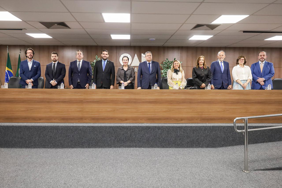 Plano médio de onze pessoas, homens e mulheres em trajes formais, alinhados lado a lado atrás de uma longa bancada de madeira em um auditório. Ao centro, na parede de madeira ao fundo, destaca-se o logotipo prateado da OAB. O ambiente é amplo, com teto de luminárias quadradas, carpetes cinza e bandeiras à esquerda.