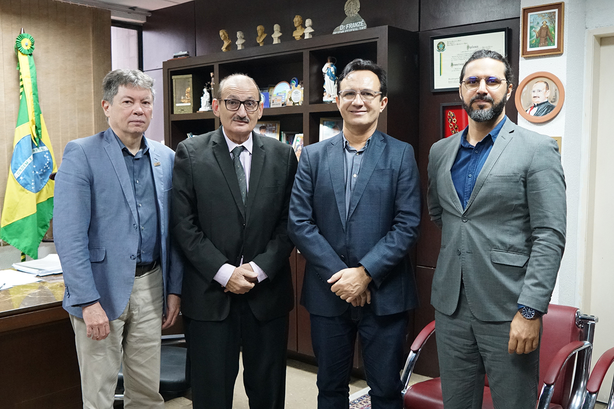 Quatro homens posam lado a lado em um escritório formal. À esquerda, a bandeira do Brasil; ao fundo, uma estante com bustos e diplomas. Eles vestem trajes sociais, alternando entre blazers cinza, preto e azul, transmitindo um ambiente de reunião institucional ou acadêmica.