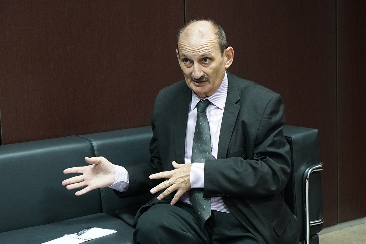 Plano médio de um homem de bigode, vestindo terno preto e gravata verde, sentado em um sofá escuro. Ele está com as mãos gesticulando à frente do corpo, como se explicasse um ponto importante durante uma conversa. Ao lado, um bloco de notas e uma caneta.