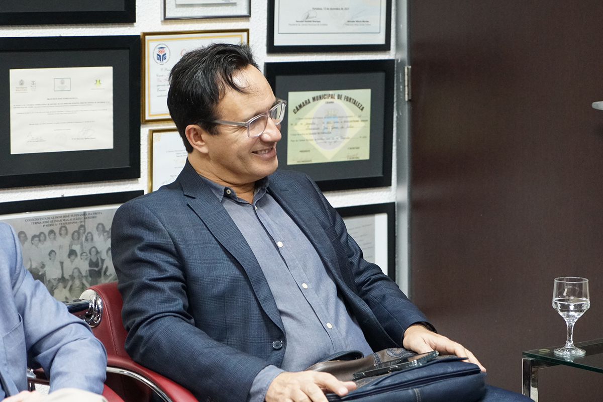 Um homem de óculos e blazer azul-marinho aparece sorrindo lateralmente, sentado em uma poltrona vermelha. Ao fundo, uma parede branca repleta de quadros e certificados emoldurados, incluindo um da Câmara Municipal de Fortaleza. Sobre uma mesa de vidro à direita, há um copo d'água.