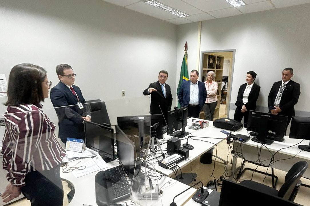 Sete pessoas estão reunidas em um escritório de paredes brancas com mesas dispostas em "L", repletas de computadores e cabos. À esquerda, em primeiro plano, uma mulher com camisa listrada e um homem de terno observam o grupo. Ao centro, um homem de terno gesticula enquanto fala, posicionado próximo a uma bandeira do Brasil. À direita, outros três homens e duas mulheres, vestidos formalmente, acompanham a conversa atentos.