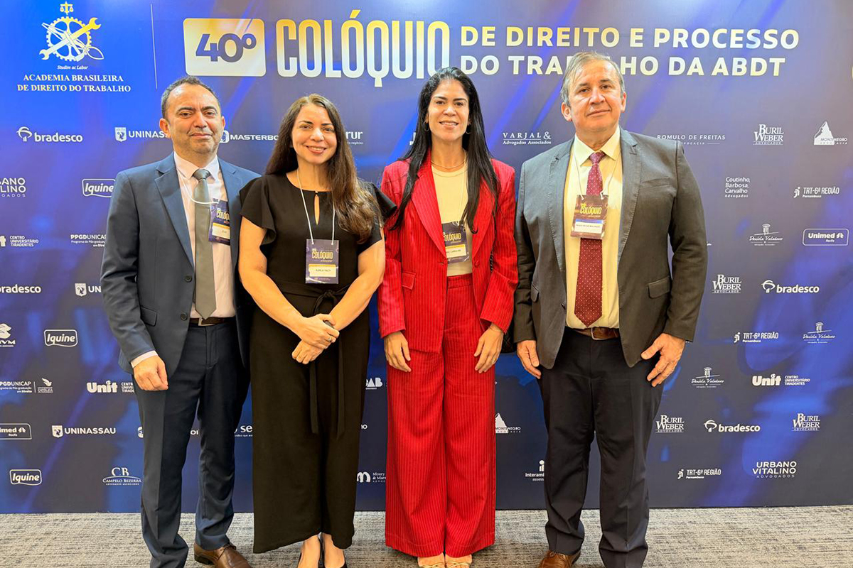 Quatro pessoas — dois homens e duas mulheres — posam alinhadas e sorridentes à frente de um painel azul institucional do "40º Colóquio de Direito e Processo do Trabalho da ABDT". Os homens vestem ternos (azul-escuro e cinza com gravata vermelha) e as mulheres usam trajes sociais, destacando-se uma delas ao centro com um terno vermelho vibrante. Todos portam credenciais oficiais do evento no pescoço, e o fundo exibe diversas logomarcas de patrocinadores em letras brancas sobre a superfície azul.