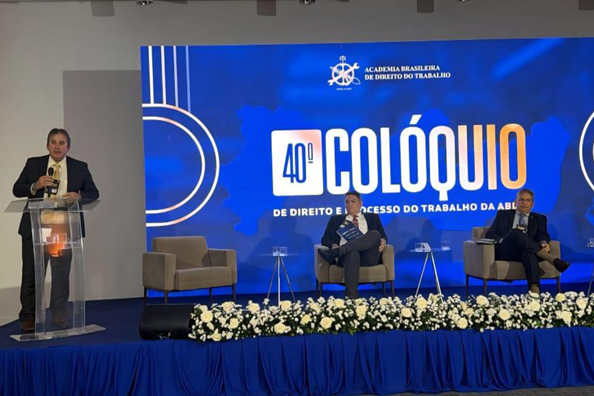 Palco de um evento formal com um grande telão azul ao fundo que exibe o logotipo do "40º Colóquio de Direito e Processo do Trabalho". À esquerda, um homem de terno fala ao microfone em um púlpito de acrílico transparente, enquanto outros dois palestrantes estão sentados em poltronas beges no centro do palco. A base da estrutura é decorada com uma longa fileira de flores brancas e folhagens, complementada por uma saia de tecido azul que cobre toda a frente do tablado.