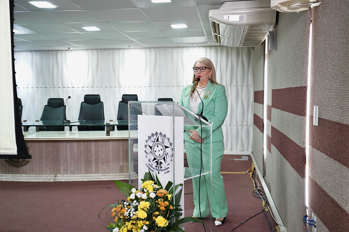 Ela está de pé atrás de um púlpito de acrílico transparente com o brasão do Brasil, falando em um microfone. Ela veste o mesmo blazer verde-água, blusa de renda branca e colar de pérolas da Imagem 1.