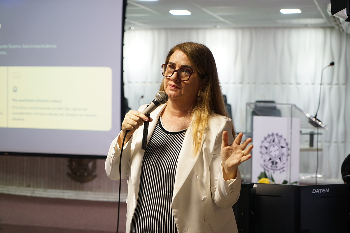 mulher de cabelos castanhos da Imagem 1: Ela está em pé, segurando um microfone com a mão esquerda e gesticulando com a mão direita. Ela veste o mesmo blazer branco sobre o vestido listrado em preto e branco 
