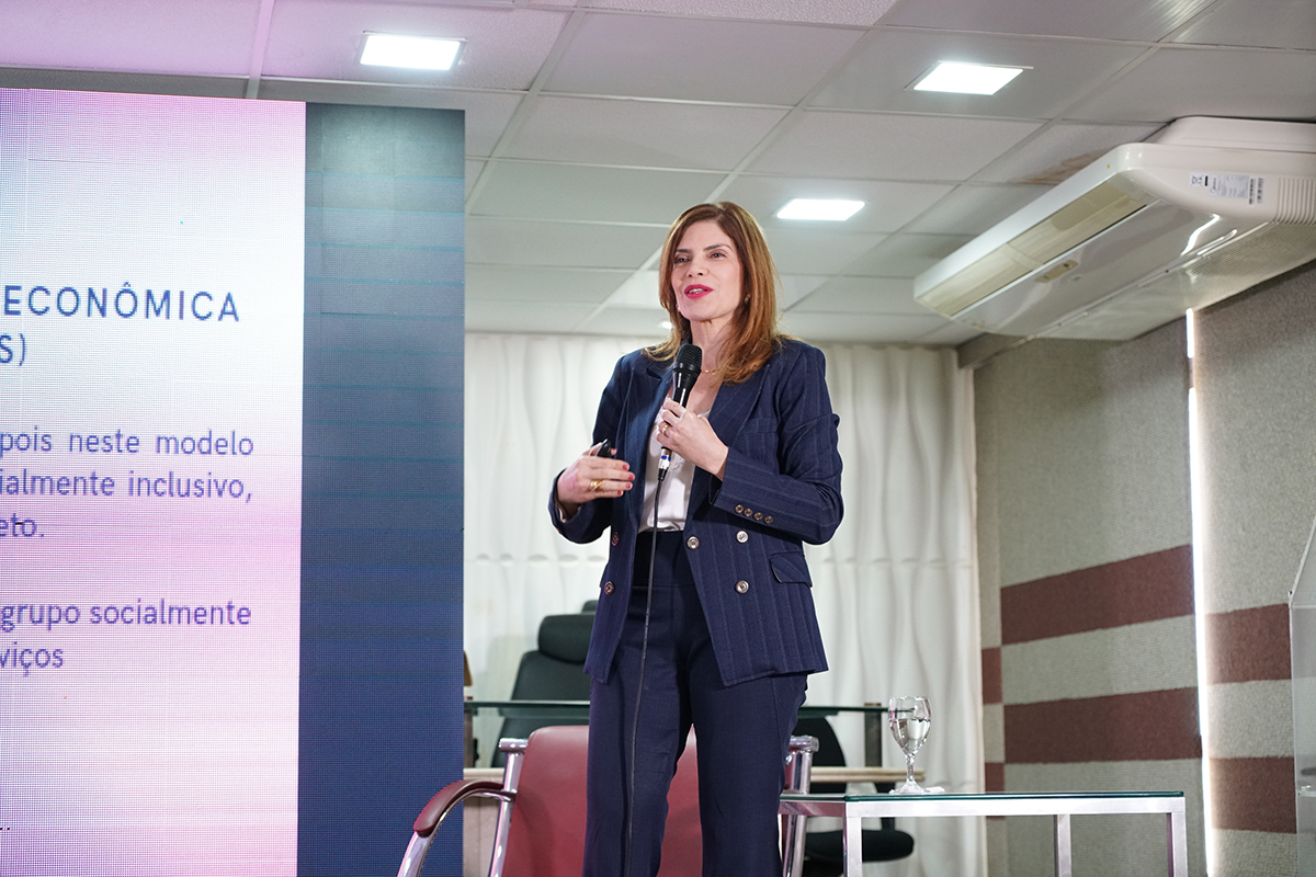 Mulher de pele clara e cabelos castanhos claros na altura dos ombros gesticula de pé no palco enquanto segura um microfone preto. Ela traja um terno risca-de-giz azul-marinho com botões dourados sobre uma blusa branca e usa batom de tonalidade vermelha destacada. À esquerda, parte de uma tela projeta tópicos textuais em azul e roxo, tendo ao fundo luminárias embutidas no teto do auditório.