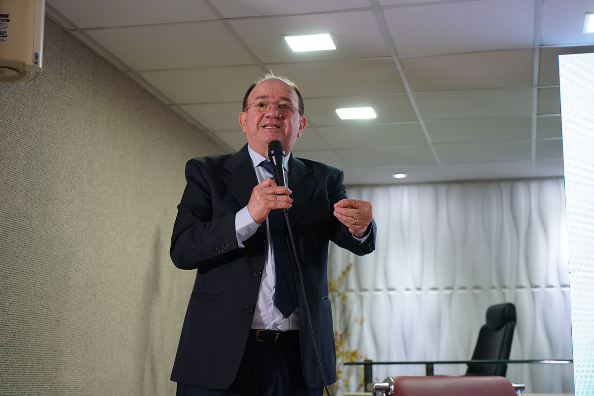 Homem sorridente de óculos e cabelos curtos discursa de pé em plano médio, vestindo terno preto, camisa azul-clara e gravata escura. Ele segura o microfone com a mão direita e mantém a mão esquerda erguida na altura do peito com os dedos levemente flexionados. O fundo apresenta uma parede de textura cinza à esquerda, luzes de LED acesas no teto e cortinas brancas ao fundo da sala.