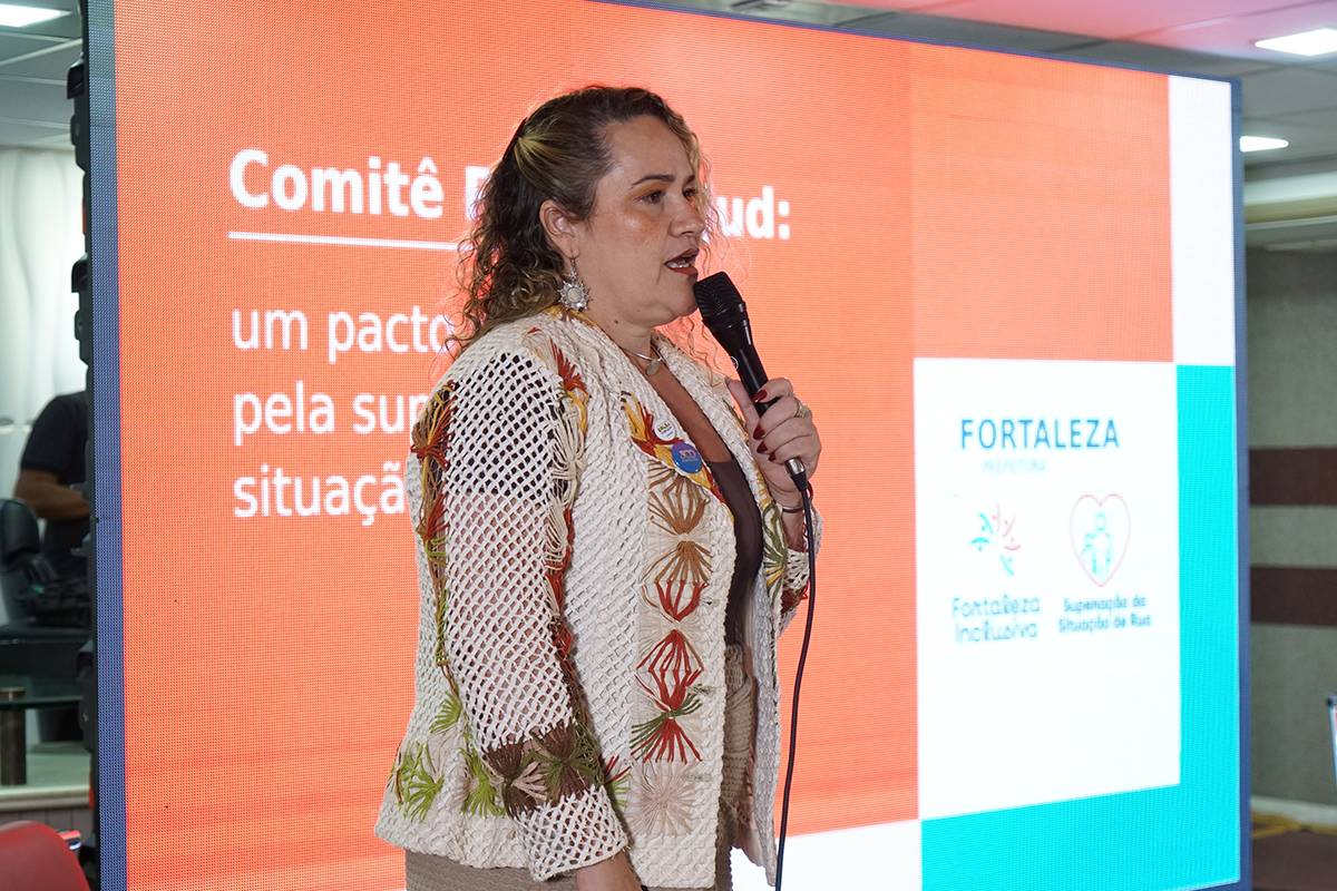 Mulher de cabelos ondulados gesticula e discursa de perfil para a direita, segurando um microfone preto próximo ao rosto. Ela veste uma blusa escura sob um casaco artesanal de trama aberta bege, decorado com bordados coloridos em relevo. Ao fundo, um painel digital com luz alaranjada destaca trechos do texto institucional "Comitê PopRuaJud" e a marca da Prefeitura de Fortaleza.