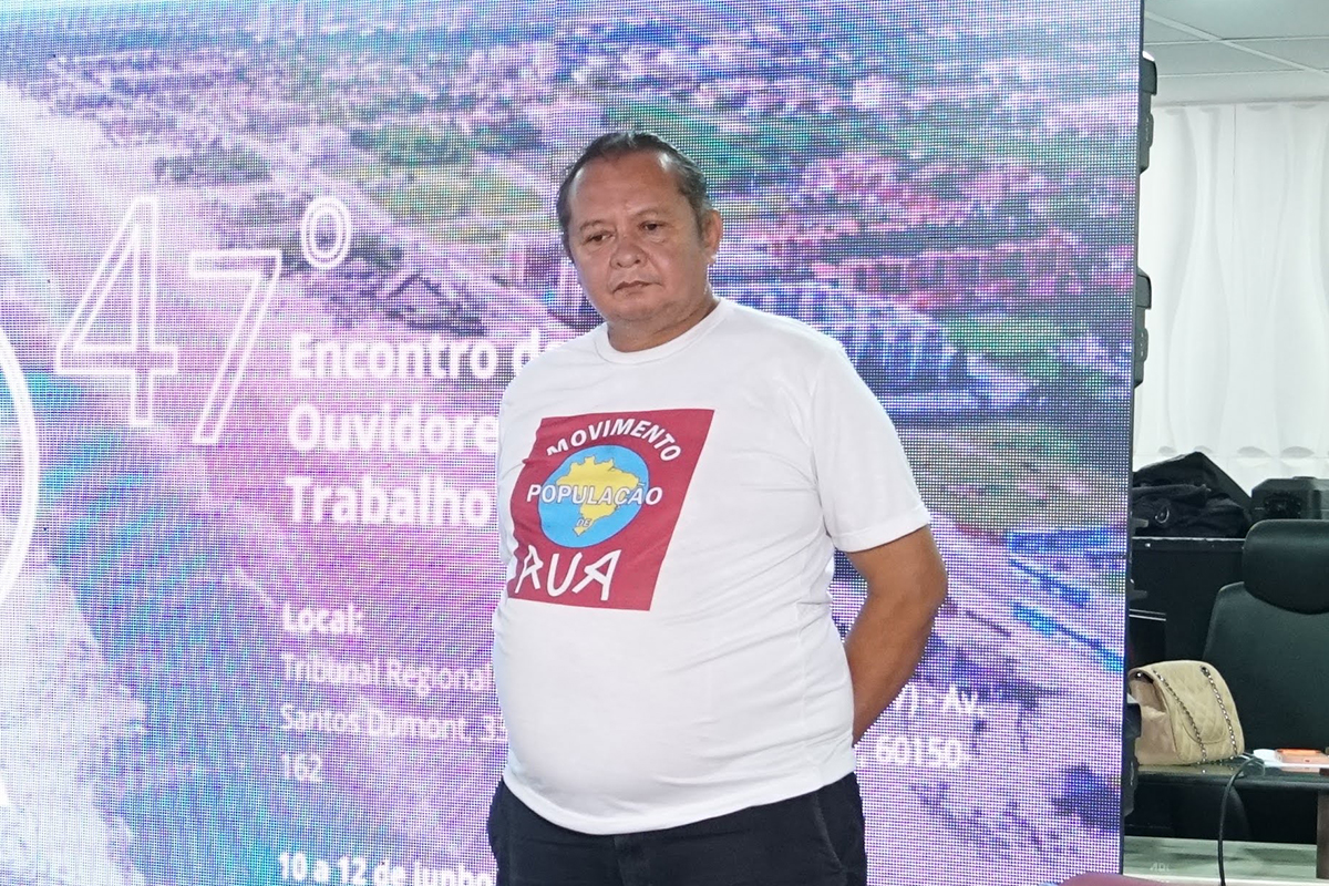 Homem de pele morena e cabelos curtos posa de pé com os braços para trás à frente de um painel digital iluminado. Ele traja uma camiseta branca com uma estampa circular central que traz o mapa do Brasil e a inscrição "Movimento População de Rua". A imagem está enquadrada em plano médio, exibindo parte do texto do evento ao fundo e uma bolsa bege sobre uma bancada lateral.