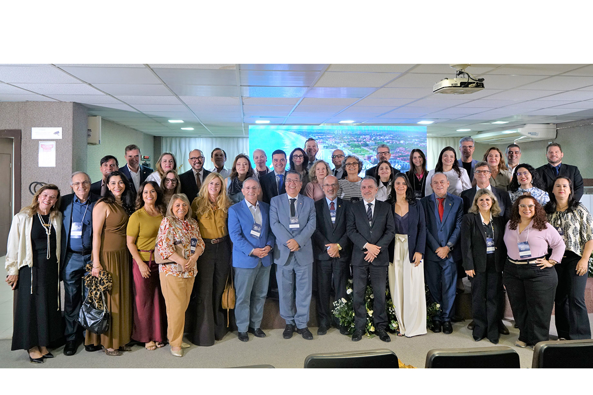 Um grande grupo de homens e mulheres posa reunido e sorridente para uma foto oficial sobre o palco do auditório. Vestindo trajes formais e casuais diversos, eles se alinham à frente de um grande telão que exibe uma imagem aérea litorânea. Na parte inferior, em primeiro plano, observam-se detalhes parciais de poltronas pretas destinadas à plateia do evento.