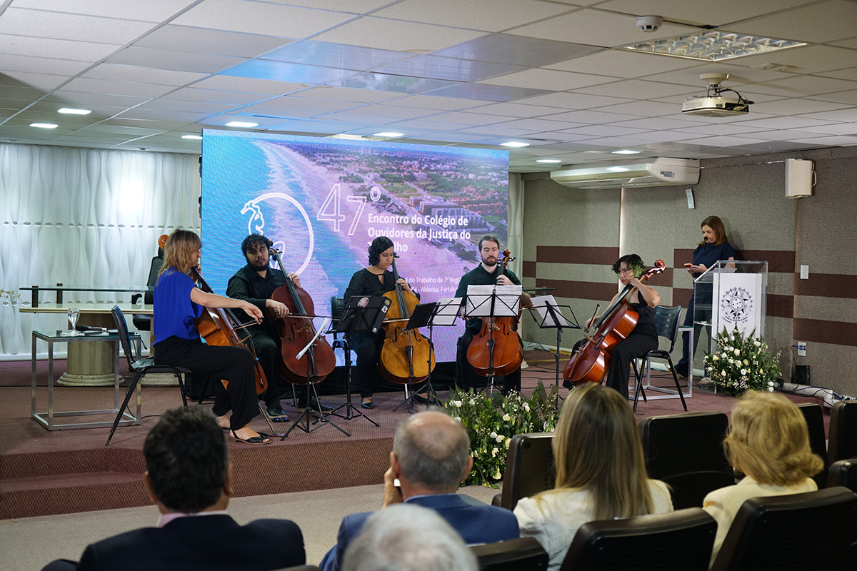 **Audiodescrição para acessibilidade:**  **Coleouv_12.6.2026_1.jpg**: No palco de um auditório, cinco musicistas (duas mulheres e três homens) sentados em semicírculo tocam violoncelo à frente de um grande telão azul iluminado com o texto do "47º Encontro". Em primeiro plano, de costas, a plateia acompanha atentamente a apresentação musical.