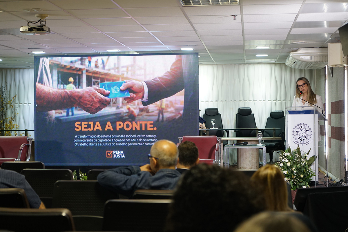 Vista do auditório focado no palco onde a juíza Izabela Ramos Pinto fala em um púlpito acrílico à direita. O telão ao centro exibe a campanha "Pena Justa", mostrando um aperto de mãos onde uma delas entrega uma carteira de trabalho.