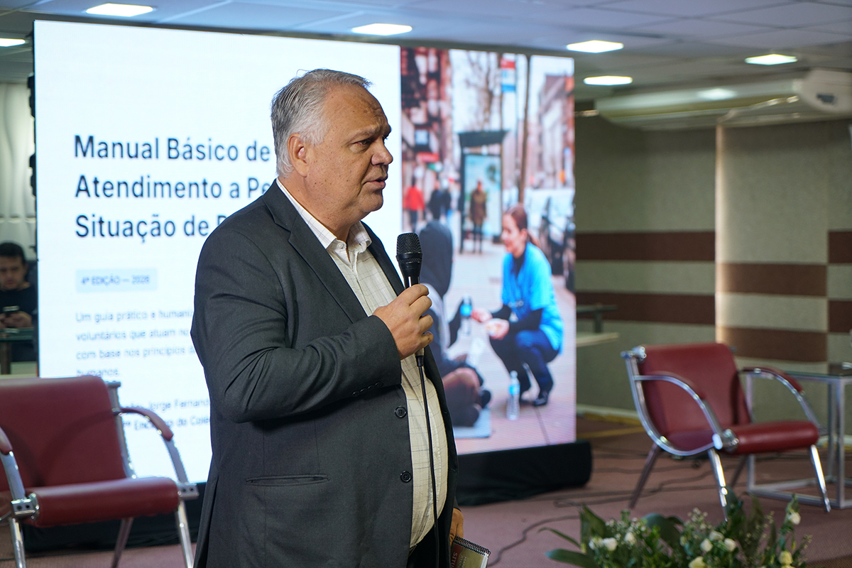 Em primeiro plano de perfil, o gestor Jorge Fernandes usa terno cinza e fala ao microfone. Ao fundo, o telão projeta o título "Manual Básico de Atendimento a Pessoas em Situação de Rua" ao lado da foto de uma abordagem social.