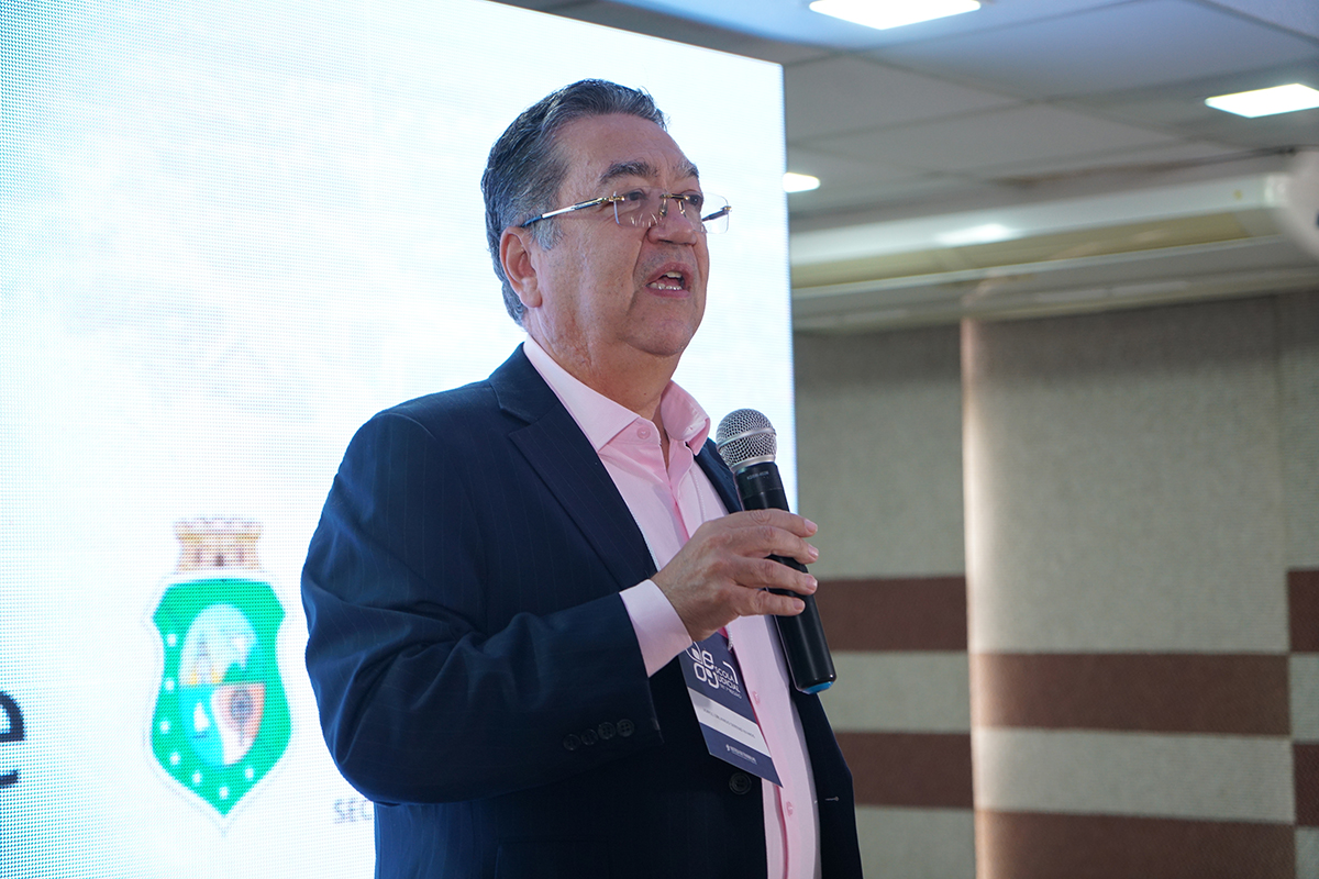 Close-up do desembargador Jorge Orlando Sereno Ramos discursando com o microfone na mão direita. Ele veste paletó escuro, camisa rosa-claro e usa óculos, posicionado em frente ao painel digital iluminado.