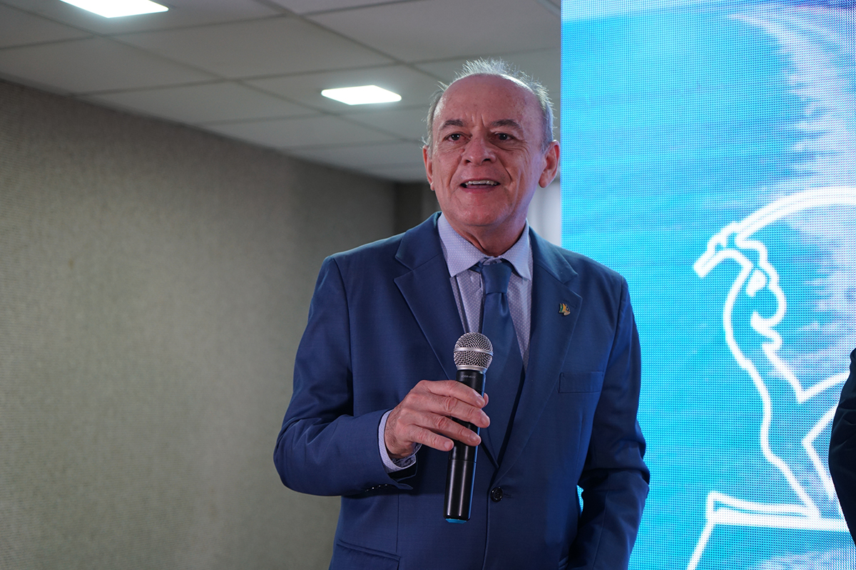 Close-up do desembargador José Antônio Parente da Silva sorrindo e falando ao microfone. Ele veste terno azul, camisa social clara e gravata azul, posicionado ao lado do luminoso do evento.