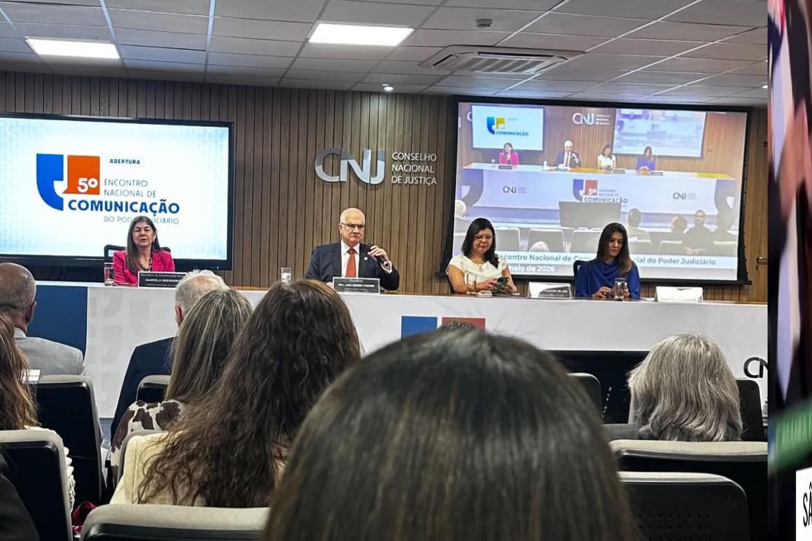 Visão a partir da plateia de um auditório focado na mesa diretora do "5º Encontro Nacional de Comunicação do Poder Judiciário", sob o logotipo do CNJ na parede de madeira. Quatro autoridades — duas mulheres nas extremidades e um homem e uma mulher ao centro — estão sentadas atrás de uma bancada branca com microfones e telas digitais ao fundo. Em primeiro plano, destaca-se a silhueta das cabeças e cabelos dos espectadores de costas.