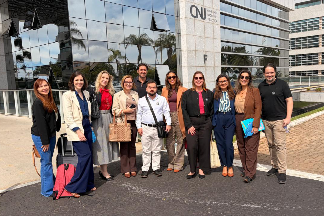 Um grupo de onze pessoas posa sorridente em uma área externa pavimentada, em frente à fachada moderna e espelhada do prédio do Conselho Nacional de Justiça (CNJ). Entre os integrantes, há homens e mulheres vestindo trajes casuais e formais, incluindo blazers, calças compridas e camisas sociais em tons variados. O reflexo dos vidros escuros do edifício exibe palmeiras e o céu com nuvens claras ao fundo.