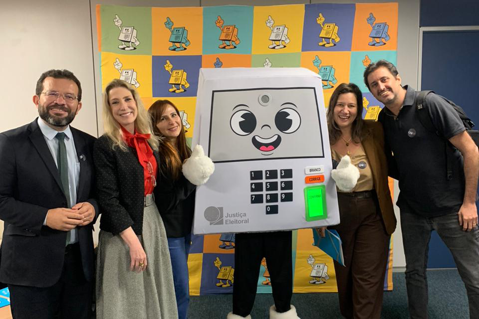 Cinco pessoas sorriem e posam abraçadas ao redor de uma mascote fantasiada de urna eletrônica com braços e pernas, que faz sinal de positivo. O grupo está em um ambiente interno, posicionado à frente de um painel quadriculado com ilustrações coloridas da própria mascote. À esquerda, um homem de terno e duas mulheres; à direita, uma mulher com blazer caramelo e um homem de camisa polo escura.