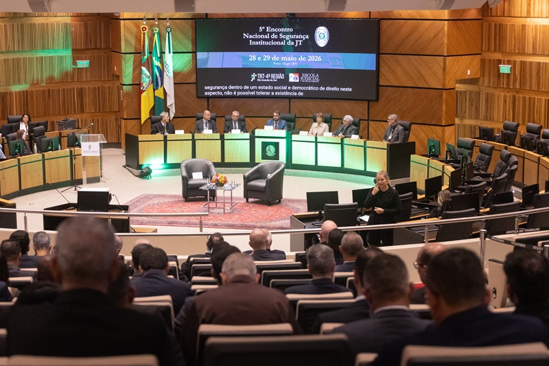 Fotografia em plano geral e perspectiva elevada mostrando um auditório durante um evento formal. No primeiro plano, de costas para a câmera, vê-se a plateia sentada em poltronas escuras. Ao fundo, no palco, autoridades estão reunidas em uma bancada semicircular, sob um grande telão digital.  Descrição Detalhada Primeiro Plano: Fileiras de poltronas de um auditório, ocupadas por diversas pessoas vistas de costas (majoritariamente homens de cabelos curtos ou grisalhos). O foco está ligeiramente desfocado nesta área, direcionando a atenção para o palco.  O Palco: No centro do palco, há duas poltronas individuais de couro cinza-escuro voltadas para a plateia, separadas por uma pequena mesa de centro de vidro com um arranjo de flores coloridas. À direita do palco, uma intérprete de Libras (Língua Brasileira de Sinais), mulher loira vestindo roupa preta, está de pé fazendo sinalizações.  A Mesa de Autoridades: Uma grande bancada de madeira clara com detalhes pretos e iluminação em LED verde na base abriga sete pessoas sentadas (cinco homens e duas mulheres), todas vestindo trajes formais. À esquerda da bancada, estão hasteadas três bandeiras oficiais em pedestais.  O Telão: Um grande painel digital exibe um fundo escuro com os textos informativos do evento:  5º Encontro Nacional de Segurança Institucional da JT 28 e 29 de maio de 2026 — Porto Alegre (RS)  Abaixo do título, constam as marcas do TRT-4ª Região e da Escola Judicial. Na parte inferior do telão, há uma faixa preta exibindo legendas em tempo real da fala das autoridades.  Ambiente: As paredes laterais e de fundo do auditório são revestidas com painéis de madeira vertical texturizada. Há galerias laterais com cadeiras de escritório vazias dispostas em bancadas.