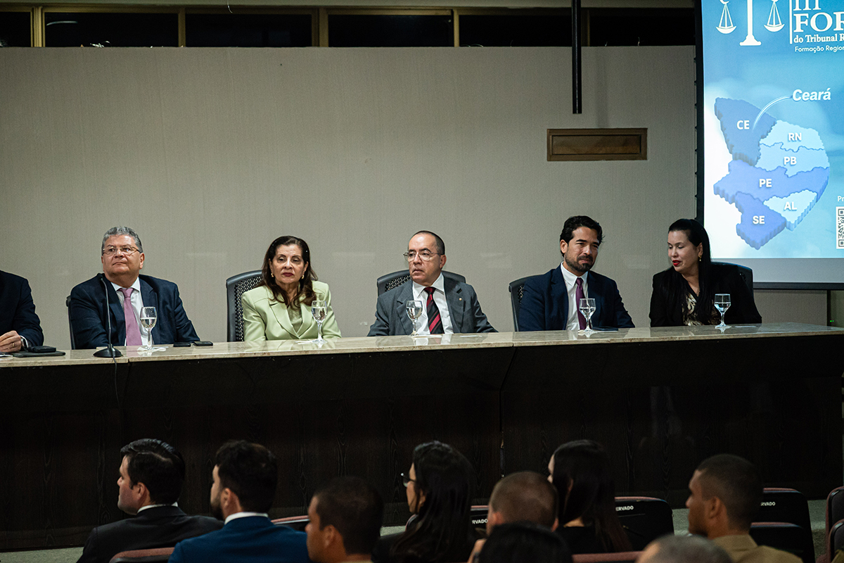 A imagem mostra o palco de um auditório onde cinco pessoas, vestidas formalmente com ternos e tailleurs, estão sentadas atrás de uma longa mesa escura com copos de água à frente. Ao fundo, uma parede clara serve de cenário, e à direita destaca-se uma tela de projeção exibindo um mapa do Nordeste brasileiro, correspondente ao arquivo Fórum Regional dos Juízes Criminais2.jpg. Em primeiro plano, vê-se a parte superior da cabeça e os ombros de algumas pessoas da plateia que assistem ao evento de costas para a câmera.
