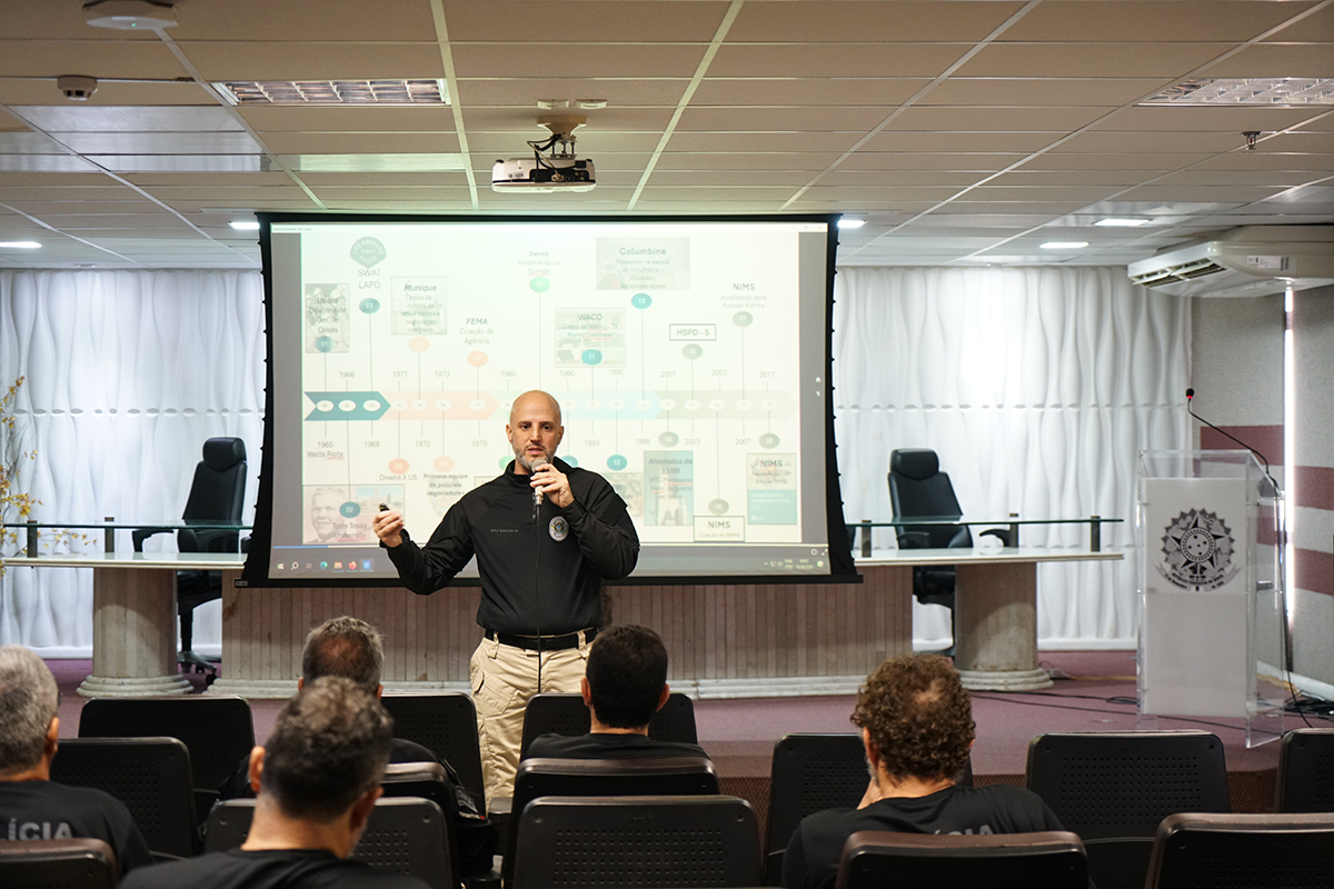 Um homem calvo vestindo camisa preta e calça cargo está de pé em um palco, falando ao microfone. Atrás dele, há uma grande tela de projeção exibindo um slide com uma linha do tempo histórica, contendo rótulos de texto e datas específicas.. Ele faz a apresentação para uma plateia sentada em cadeiras escuras, em um auditório moderno e bem iluminado