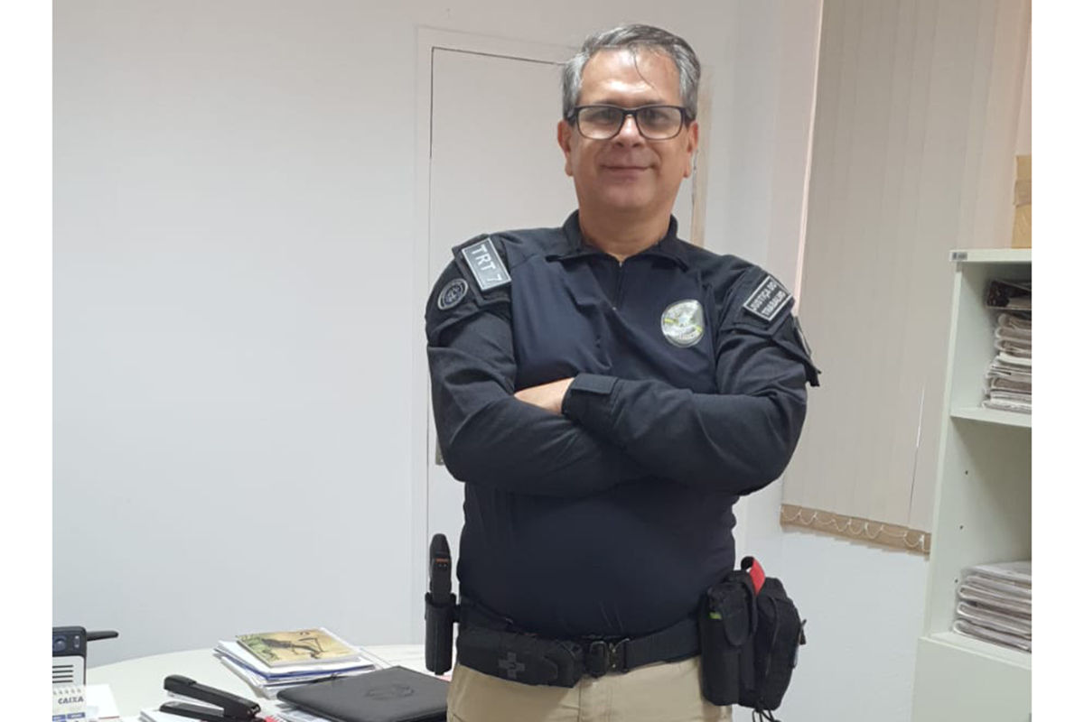 Um homem de meia-idade com óculos e cabelos grisalhos, vestindo farda preta do TRT7 e cinto tático, está de pé em um escritório com os braços cruzados, sorrindo. 