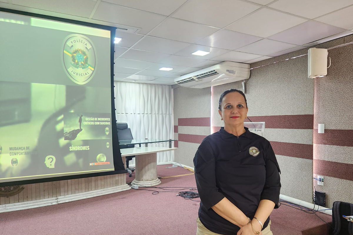 Uma mulher de pele clara, cabelos escuros presos e usando batom vermelho está em pé, em primeiro plano e ligeiramente à direita, posicionada em um auditório. Ela veste uma camisa tática preta de mangas três quartos com o brasão da Polícia Judicial no lado esquerdo do peito e calça bege clara, mantendo as mãos unidas à frente do corpo. À esquerda, ao fundo, destaca-se um grande telão com projeção esverdeada exibindo o brasão oficial da Polícia Judicial e os títulos "Gestão de Incidentes Críticos com Suicidas", "Síndromes" e "Mudança de Comportamento". O ambiente do auditório possui piso acarpetado avermelhado, teto de placas brancas com luminárias embutidas, paredes texturizadas em tons de cinza e marrom com um aparelho de ar-condicionado fixado, além de uma mesa de vidro com poltrona preta posicionada logo atrás do telão.