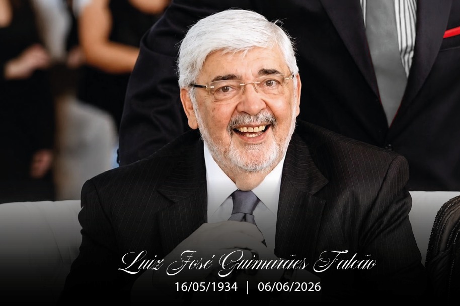 Retrato de um homem idoso sorridente, de pele clara, cabelos brancos curtos, barba rala grisalha e óculos de grau com armação fina, vestindo terno preto listrado, camisa branca e gravata cinza.  Ele está posicionado em primeiro plano e, ao fundo, vê-se parcialmente a silhueta de outra pessoa vestindo terno escuro com gravata cinza e lenço vermelho no bolso.  Na parte inferior da imagem, há uma faixa preta translúcida com o nome "Luiz José Guimarães Falcão" escrito em letras cursivas brancas, seguido pelas datas "16/05/1934 | 06/06/2026".