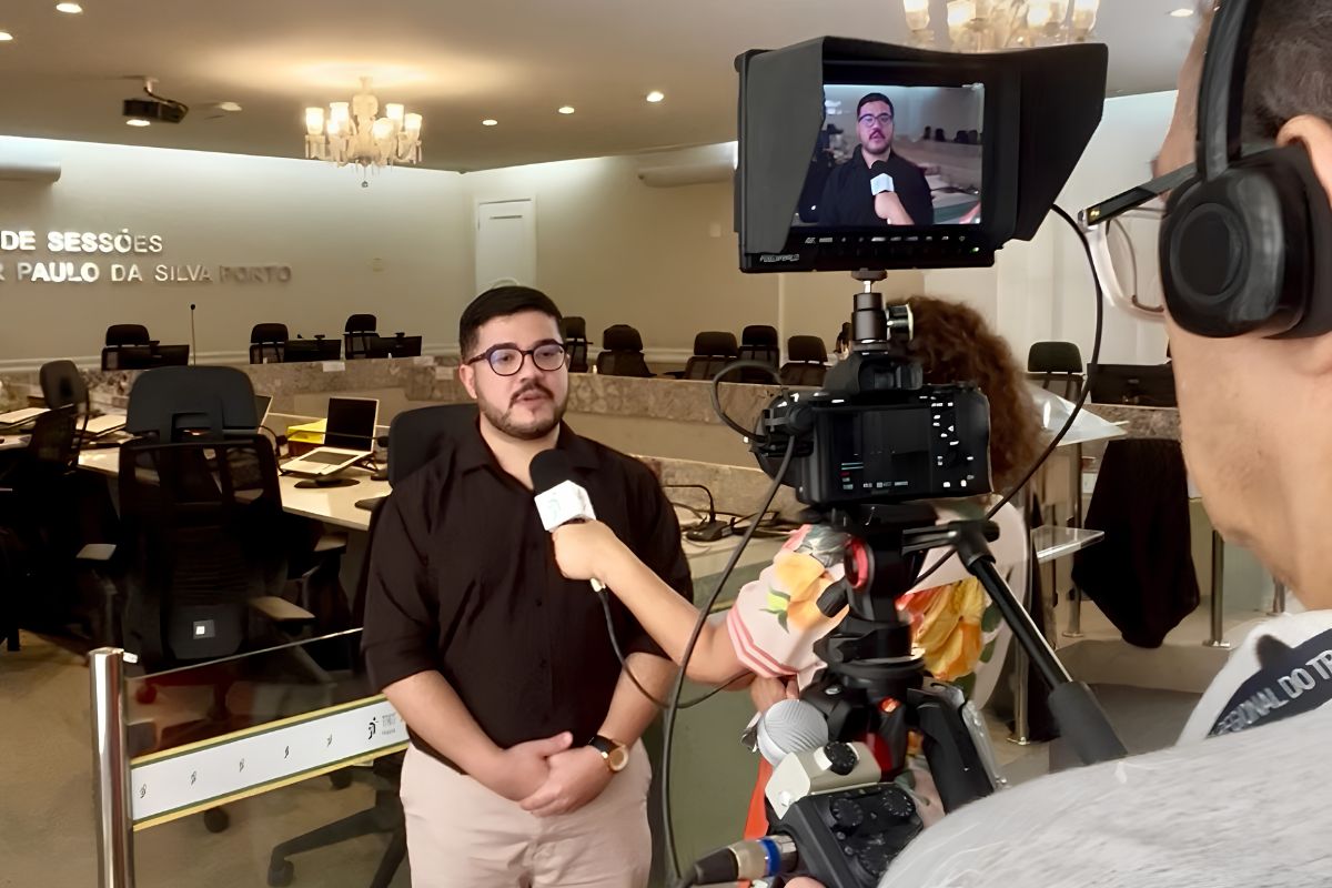 Na imagem, um homem de óculos e camisa preta concede uma entrevista em um plenário, segurando ou falando próximo a um microfone estendido por uma repórter parcial de costas. No primeiro plano, à direita, um cinegrafista com fones de ouvido opera uma câmera profissional montada em um tripé, cujo monitor exibe o enquadramento do entrevistado. Ao fundo, o amplo salão de sessões conta com mesas, cadeiras de escritório pretas