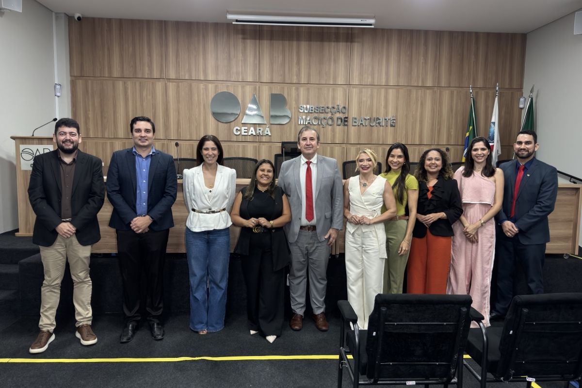 A imagem mostra dez pessoas enfileiradas e sorrindo em um auditório formal, posicionadas à frente de uma bancada de madeira elevada. Ao fundo, uma parede divisória de madeira exibe o letreiro metálico "OAB CEARÁ SUBSECÇÃO MACIÇO DE BATURITÉ", ladeado por bandeiras oficiais à direita.