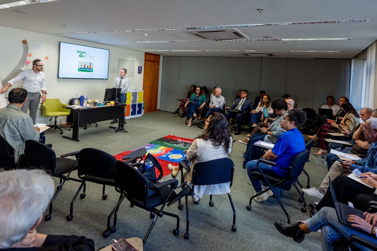 Uma sala de reuniões ampla abriga um público atento sentado em semicírculo diante de dois palestrantes posicionados à esquerda, um de pé gesticulando e outro ao lado de uma mesa com notebook e TV. No centro do tapete cinza, estende-se uma colcha colorida com inscrições. Os participantes, diversos em gênero e etnia, vestem roupas predominantemente casuais e formais, acompanhando a apresentação.