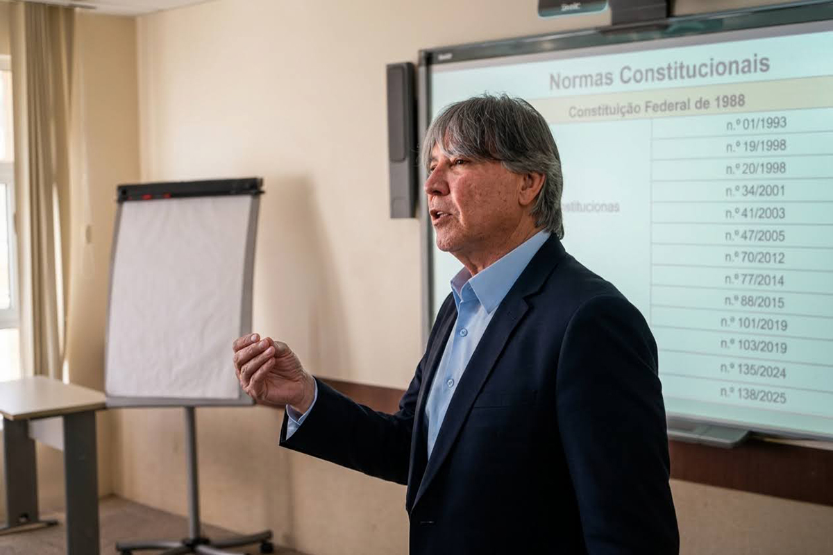 Plano médio aproximado de perfil do instrutor, um homem de pele clara, cabelos grisalhos e camisa azul sob paletó escuro. Ele gesticula com a mão direita erguida enquanto fala, expressando dinamismo e autoridade técnica. Ao fundo, de forma levemente desfocada, destacam-se um quadro branco de apoio e a tela com dados previdenciários.