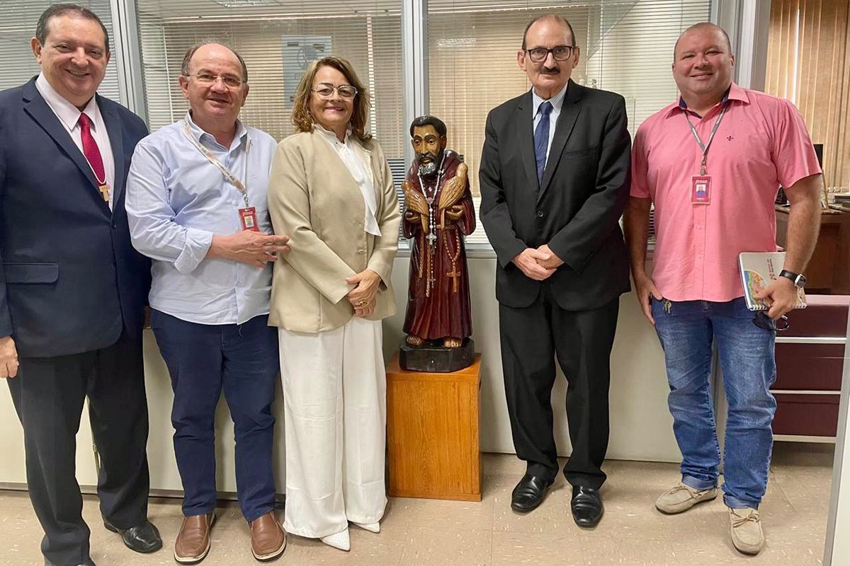A imagem Reunião_Franzé_Gomes.jpg mostra cinco pessoas, sendo quatro homens e uma mulher, posando lado a lado em pé em um escritório, sorrindo para a câmera.  Centralizada no grupo, há uma imagem religiosa de São Francisco de Assis, esculpida em madeira, posicionada sobre um pedestal do mesmo material.  O ambiente de fundo é um escritório formal com divisórias de vidro e persianas, além de armários e uma cadeira de escritório visíveis à direita.