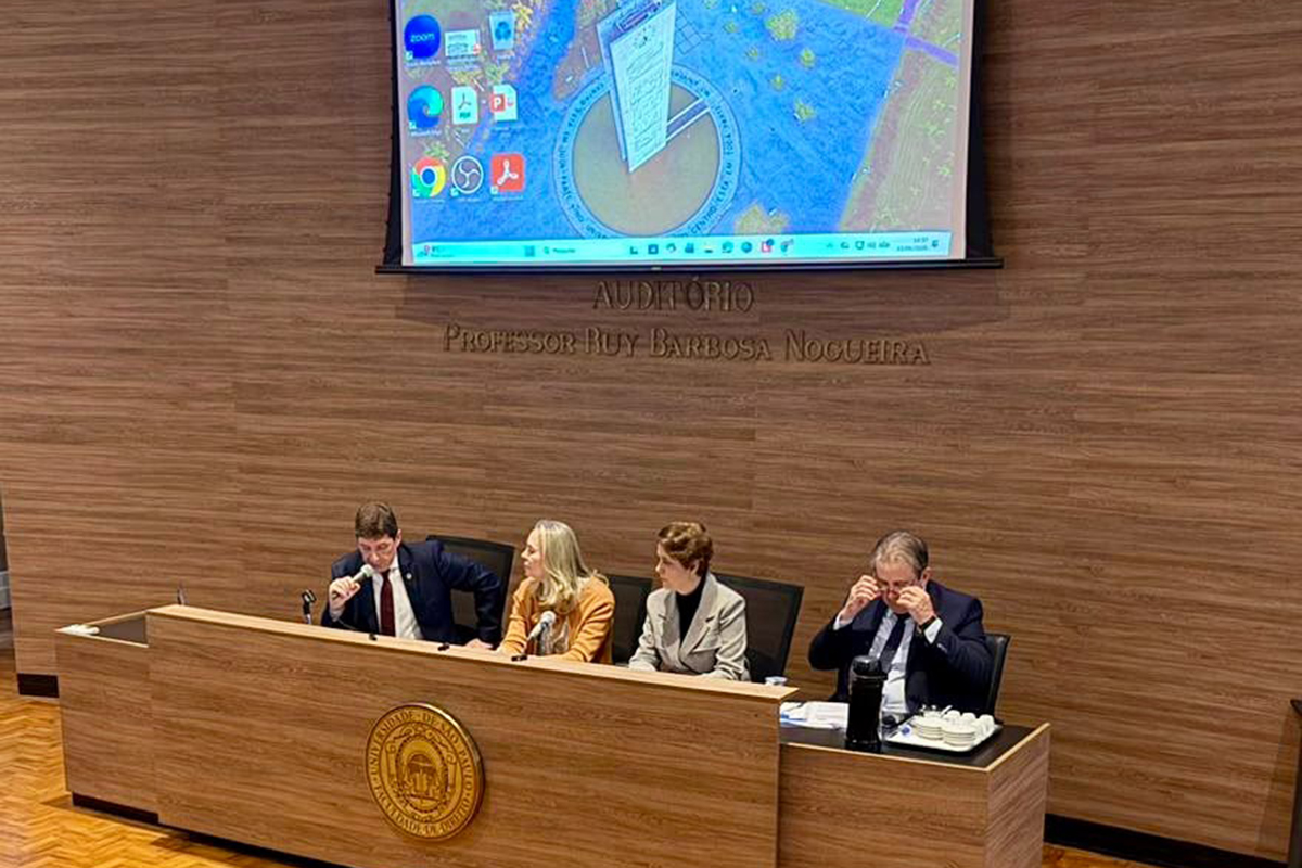 Visão elevada de uma mesa de debate composta por quatro integrantes sentados em cadeiras pretas atrás de uma bancada de madeira. Na parede de fundo texturizada, uma grande tela de projeção exibe a imagem de um slide acima do letreiro dourado do auditório. O homem posicionado na extremidade direita ajusta seus óculos enquanto os demais acompanham a fala do primeiro palestrante.