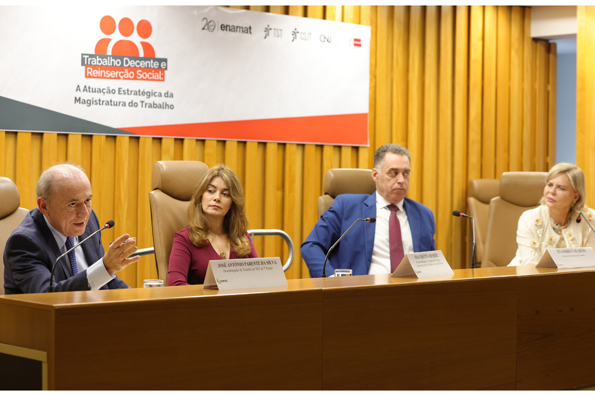 Fotografia em plano médio mostra quatro pessoas sentadas em uma mesa de conferência de madeira, posicionadas diante de uma parede com painéis verticais dourados. À esquerda, um homem calvo de terno gesticula ao falar ao microfone, ladeado por uma mulher de vestido vinho, um homem de terno azul e gravata vermelha, e uma mulher loira de blusa clara na ponta direita. Acima deles, um grande banner branco exibe o logotipo e o texto "Trabalho Decente e Reinserção Social: A Atuação Estratégica da Magistratura do Trabalho".