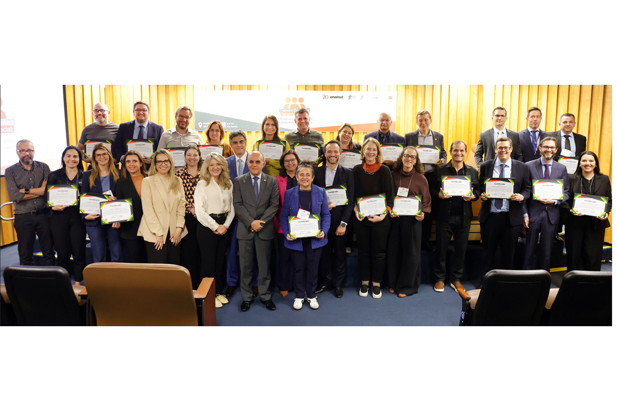 Fotografia em plano geral mostra um grupo de cerca de trinta pessoas sorridentes, posando juntas no palco de um auditório com fundo de painéis de madeira dourada. A maioria das pessoas veste trajes formais e segura certificados horizontais com bordas coloridas, voltados para a frente. O grupo está organizado em duas fileiras, com os rostos visíveis, e à frente deles aparecem as partes superiores de poltronas pretas e marrons da plateia.