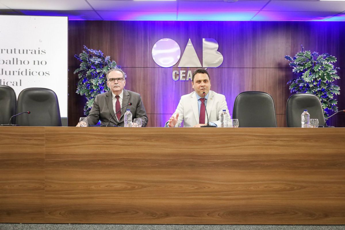 Dois homens de terno estão sentados lado a lado atrás de uma longa bancada de madeira escura em um auditório. O homem à esquerda veste terno escuro e óculos, enquanto o da direita usa terno claro e gesticula com a mão. Ao fundo, uma parede de madeira exibe o logotipo metálico iluminado da "OAB CEARÁ", ladeado por duas plantas ornamentais e luzes azuis e roxas. À esquerda, há um telão com textos em preto e, sobre a bancada, garrafas de água, copos e microfones de mesa.