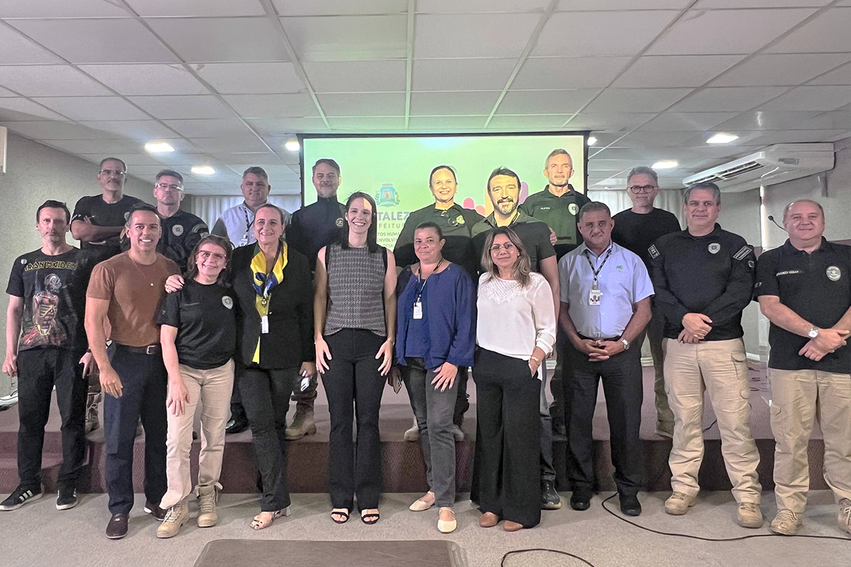 Grupo de 16 pessoas posa para foto sobre um palco em auditório. À frente, um telão projeta a logo da Prefeitura de Fortaleza e do Centro de Referência LGBT. Os integrantes — homens e mulheres de várias idades — vestem roupas casuais, camisas polo, fardamentos e calças de tons cáqui e preto, sorrindo para a câmera.