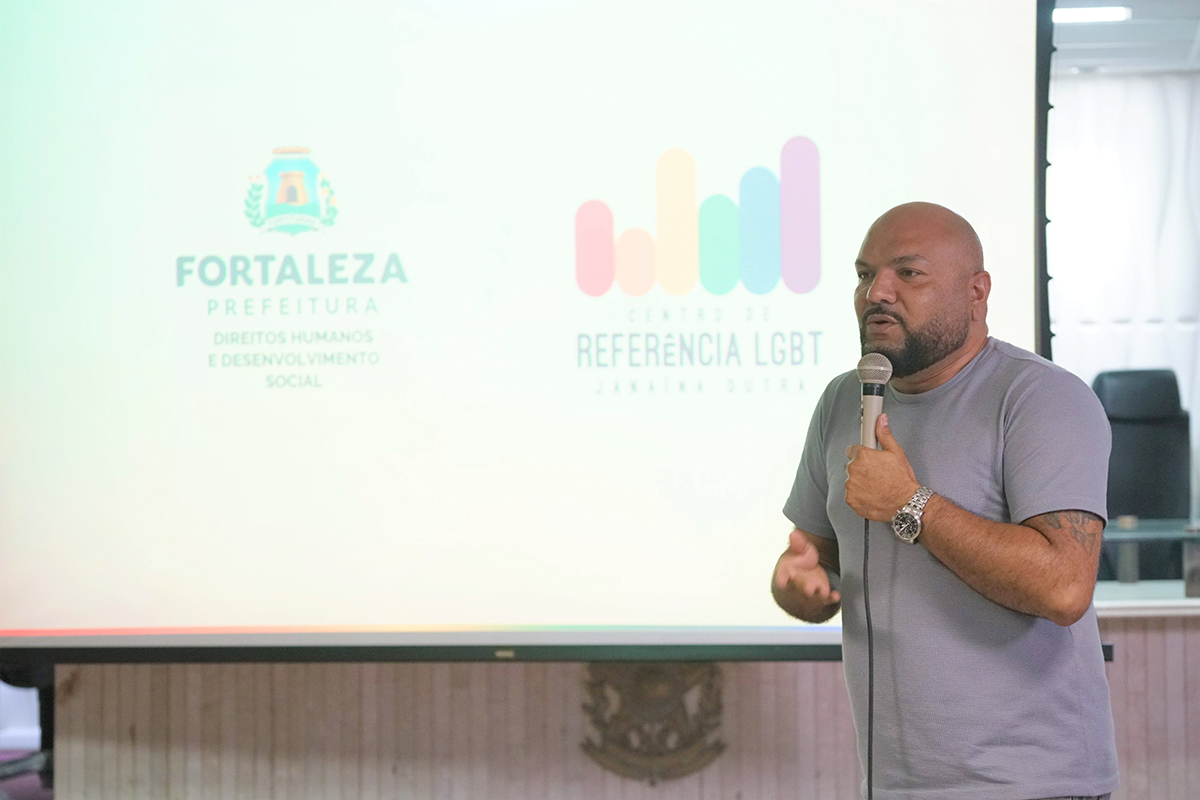 Homem pardo, calvo e com barba aparada, fala ao microfone voltado levemente para a esquerda. Ele usa uma camiseta cinza básica e relógio de pulso prateado, revelando uma tatuagem no braço esquerdo. Ao fundo, a mesma projeção clara destaca a identidade visual do Centro de Referência LGBT de Fortaleza.