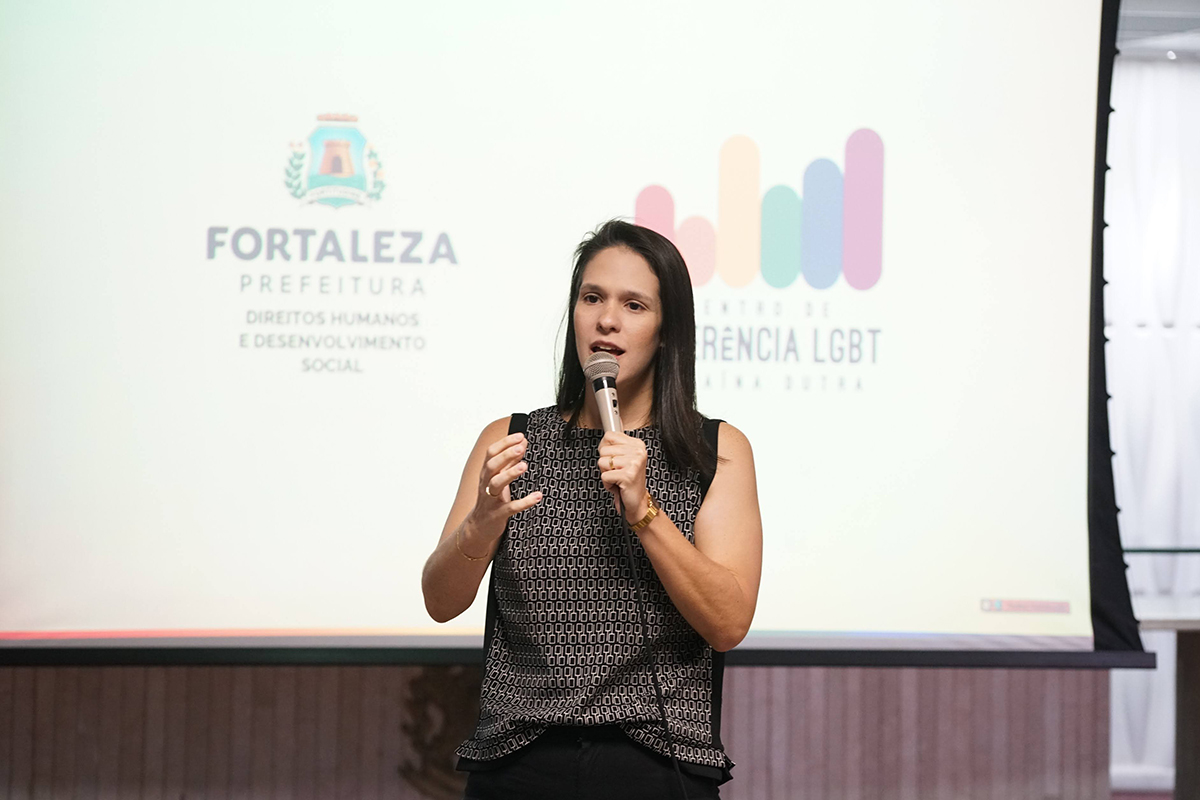 Mulher jovem, de pele clara e longos cabelos pretos lisos, fala ao microfone em um auditório. Ela veste uma blusa regata preta com estampa quadriculada cinza na frente. Ao fundo, um telão iluminado exibe o brasão da Prefeitura de Fortaleza e o texto "Centro de Referência LGBT Janaína Dutra".