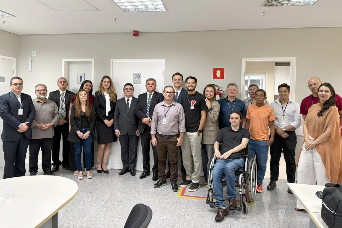 Grupo de dezenove pessoas posa para foto em uma sala de escritório de paredes claras. Elas estão dispostas lado a lado em uma linha ligeiramente curva, vestindo trajes formais e casuais variados; no centro-direita, um homem de camiseta preta e calça jeans sorri sentado em uma cadeira de rodas. À frente, nos cantos esquerdo e direito, veem-se partes de mesas brancas, e ao fundo há três portas e um extintor de incêndio na parede. 