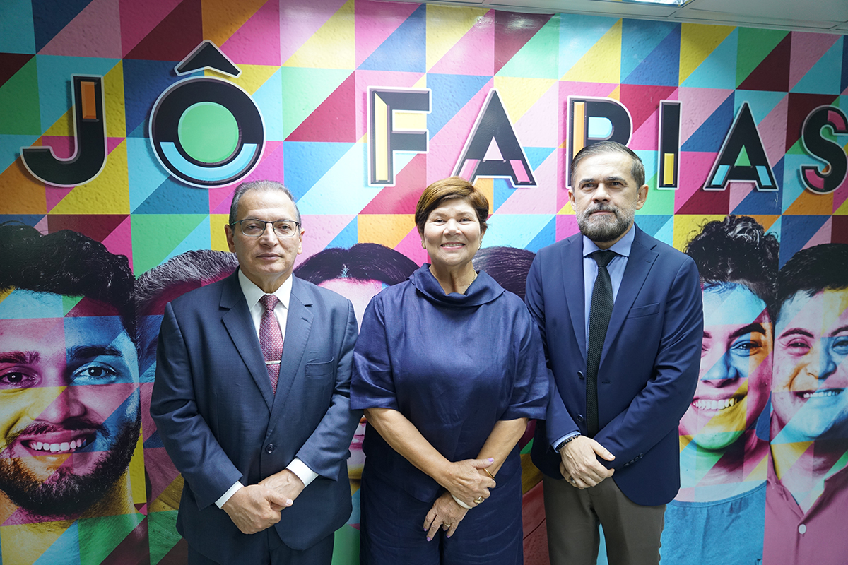 três integrantes do grupo — uma mulher ao centro vestindo blusa azul-escura e dois homens de terno e gravata — posam sorridentes lado a lado. Eles estão posicionados à frente de um painel multicolorido com formas geométricas e rostos ilustrados, onde se destaca o nome "JÔ FARIAS" em letras estilizadas no topo.