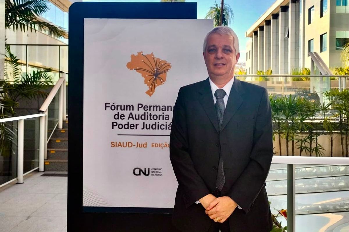 Homem de pele clara e cabelos curtos grisalhos veste terno escuro, camisa branca e gravata cinza, posando de mãos dadas à frente do corpo. Ele está posicionado em pé ao lado de um painel vertical branco com o logotipo do mapa do Brasil e a inscrição "Fórum Permanente de Auditoria do Poder Judiciário", além do logo do CNJ. Ao fundo, vê-se um ambiente externo moderno com escadaria, guarda-corpo de vidro, plantas ornamentais e a fachada de um prédio sob iluminação natural.