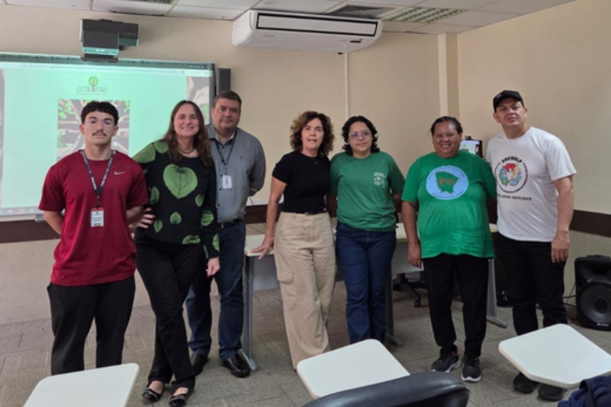 Fotografia em ambiente interno mostra um grupo de sete pessoas, quatro mulheres e três homens, posicionados lado a lado em uma sala de reuniões. À esquerda, pessoas vestem roupas casuais e formais; ao centro e à direita, integrantes usam camisetas verdes e brancas com logotipos de associações. Ao fundo, vê-se uma tela interativa de projeção, um ar-condicionado na parede e, em primeiro plano, o encosto de cadeiras escolares.