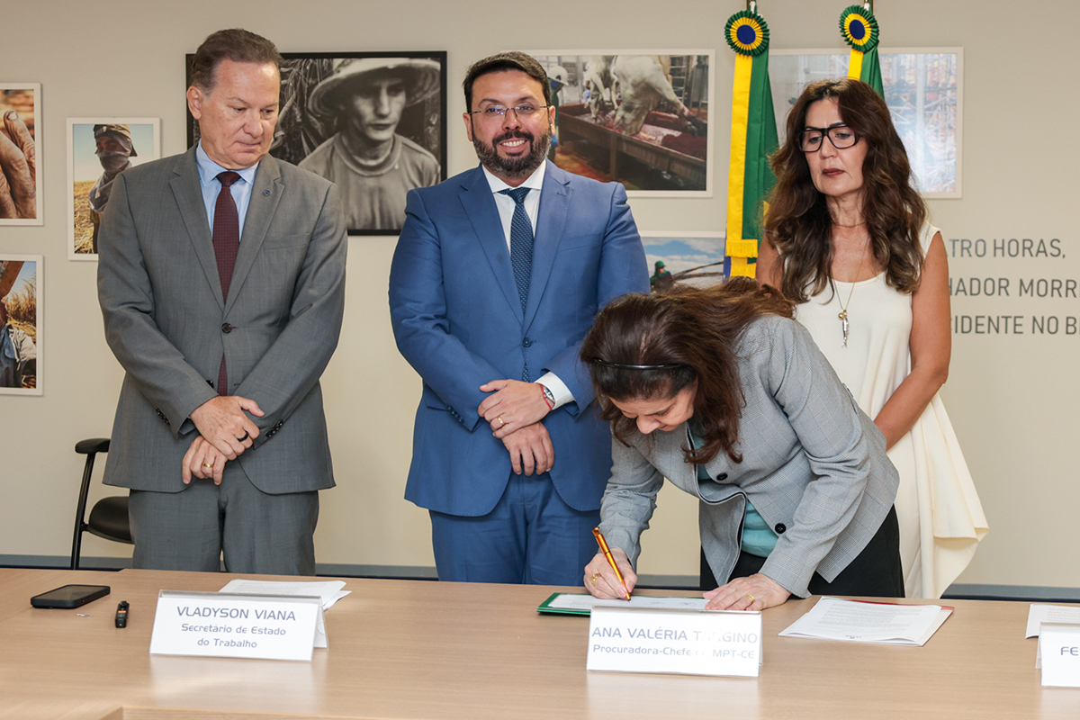 Quatro autoridades estão de pé atrás de uma mesa de madeira clara em um ambiente institucional com fotografias nas paredes. Uma mulher de blazer cinza e cabelos castanhos está inclinada, assinando um documento com uma caneta de ponta dourada sobre a mesa. Ao seu lado esquerdo, dois homens de terno (um cinza e um azul) e uma mulher de blusa branca observam atentamente o ato de assinatura.
