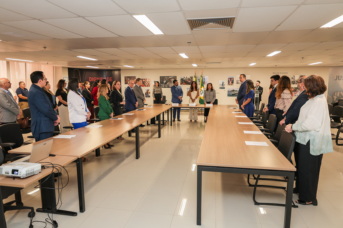 Plano geral de uma ampla sala de reuniões com formato em "U", onde dezenas de pessoas vestidas formalmente estão de pé ao redor das grandes mesas de madeira. No centro ao fundo, três autoridades permanecem posicionadas diante das bandeiras oficiais em um momento de solenidade institucional. O espaço possui iluminação embutida no teto, piso claro e paredes decoradas com quadros fotográficos.