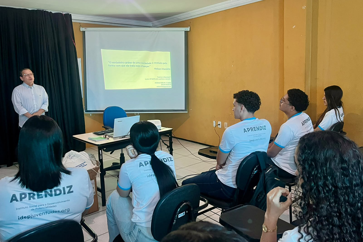 A imagem mostra a perspectiva de trás da sala, focando em jovens de cabelos escuros sentados e assistindo à apresentação com atenção. O palestrante de camisa branca está posicionado no canto esquerdo, ao lado da cortina preta e da mesa de apoio com o notebook. A tela centralizada projeta o texto verde com a frase de Nelson Mandela, iluminando suavemente o ambiente.